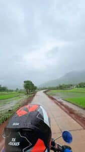 Rider in helmet on a motorcycle riding through a rain-soaked countryside road with green fields and misty hills, heading to a monsoon trek starting point
